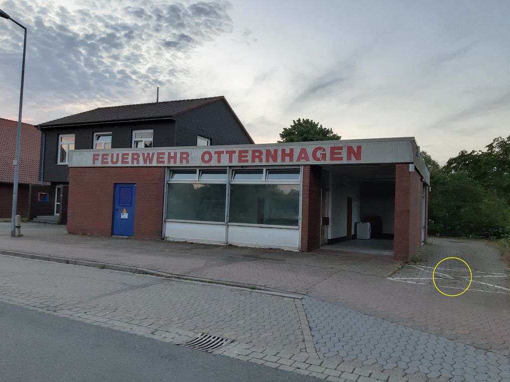 Das Bild zeigt einen vorher-nachher-Vergleich: Auf dieser Aufnahme vom 20. Juni 2019 befindet sich rechts neben dem alten Feuerwehrgerätehaus eine mit weißen Linien markierte Sperrfläche. Im Mittelpunkt dieser Sperrfläche befindet sich ein Kanaldeckel. (Gelb markiert)